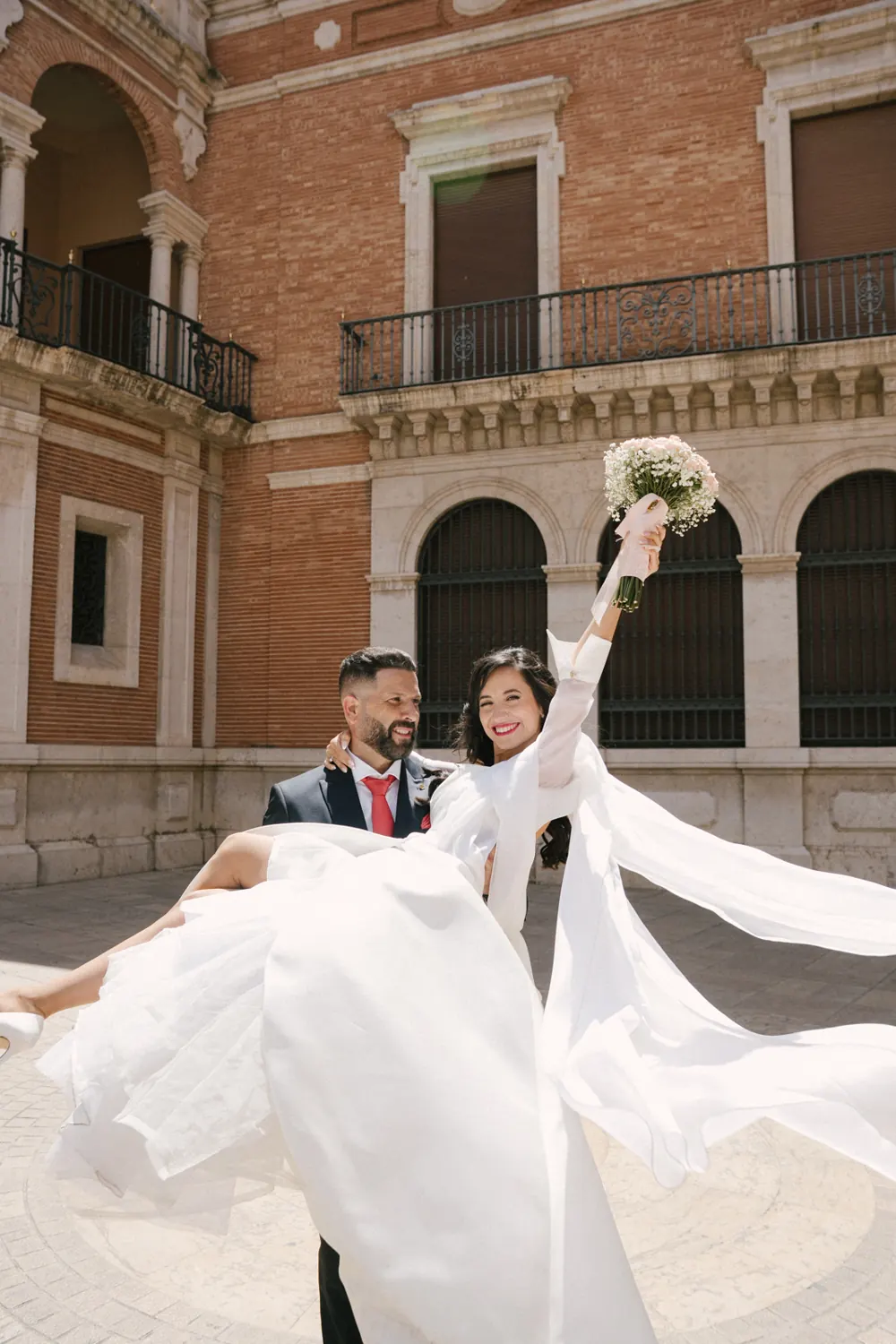 El novio coge en brazos a la novia, que levanta el ramo frente a un palacio