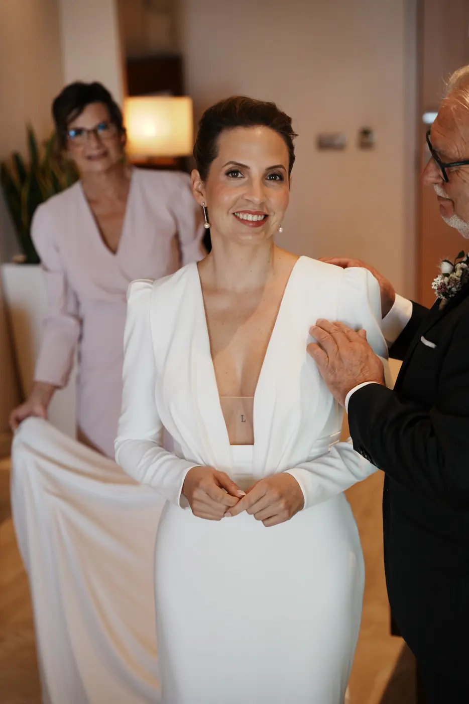 La novia sonríe mientras sus padres la ayudan con el vestido